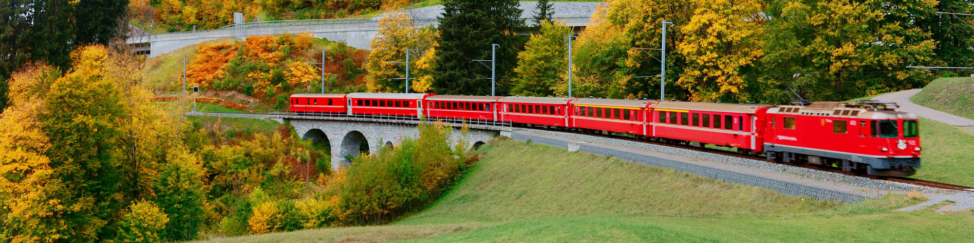 A local train of Rhaetian Railway (RhB) travels through a viaduct onto green grassy meadows with beautiful fall colors on the hillside, in Klosters village near Davos in Grisons (Graubunden), Switzerland